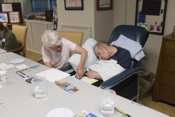 A senior adult woman helps a senior adult man with his art project