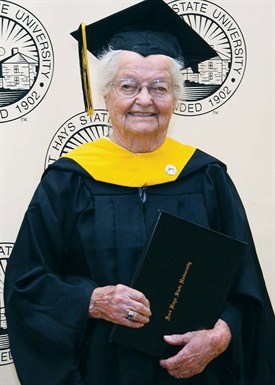 A senior adult dressed in graduation regalia and holds a diploma from Fort Hays State University