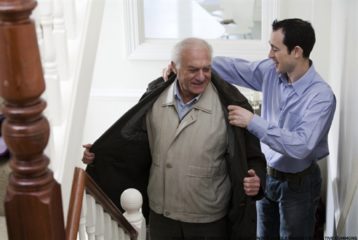 A senior adult male is helped with putting his coat on by a younger man