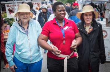 A caregiver holds the hands of two senior residents as they navigate a bustling farmers market