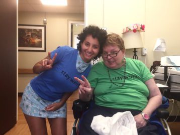 A young women and an older woman in a wheel chair. Both pose for a photo, throwing the "peace sign" with their right hands and both wearing a shirt with text across the chest that reads: "stay weird"