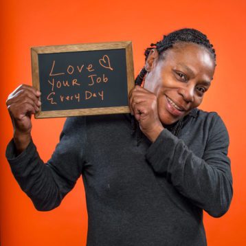 A person stands in front of an orange backdrop holding a blackboard with the handwritten text: "Love Your Job Every Day"