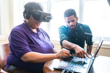 A woman wears a set of virtual reality goggles while a man next to her gives instruction