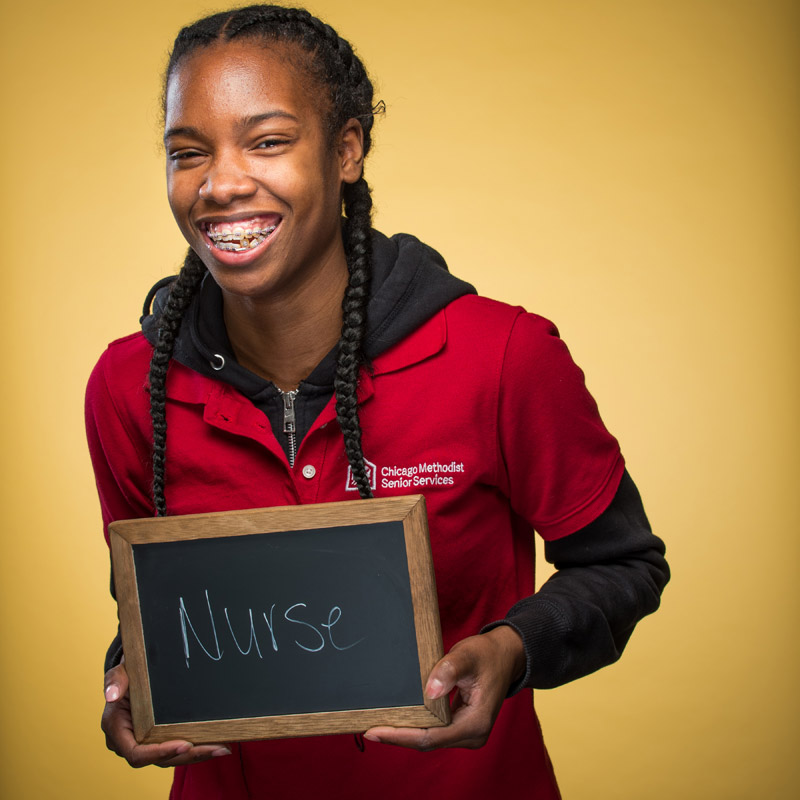 A young person stands in front of a yellow backdrop while holding a small chalkboard bearing the text: "Nurse"