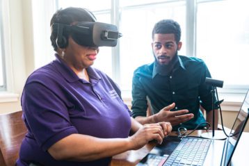 A woman wears a set of virtual reality goggles while a man next to her gives instruction