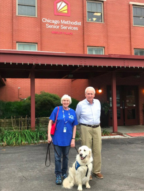 An elderly couple stand outside Wesley Place building (a multi-story brick building under operation of Chicago Methodist Senior Services) with their Golden Retriever comfort dog