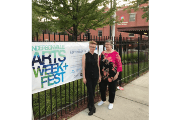 Two people stand outside alongside a banner promoting the Andersonville Arts Week & Fest