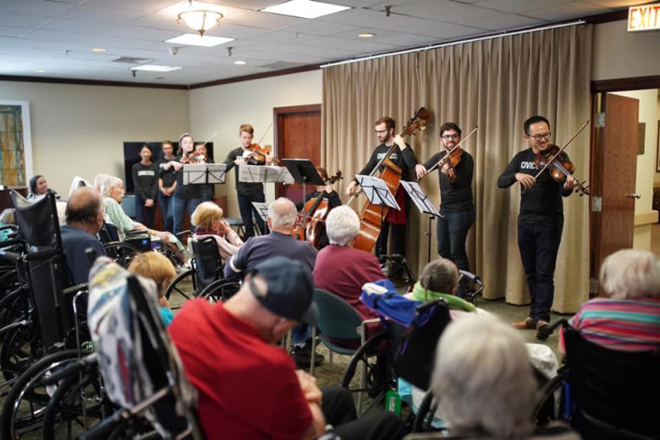 A group of musicians perform with stringed instruments in front of an audience of seniors in a common area of a residential care facility