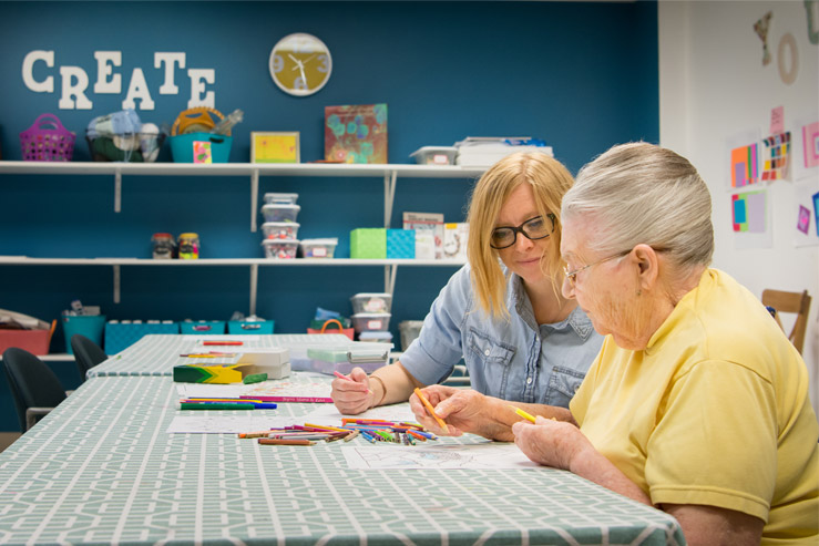 Two women sit at a table, working together on a coloring project inside an arts and crafts room.