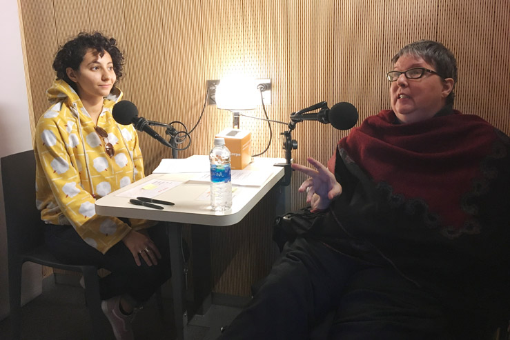 A young woman and an older woman sit together in a recording booth engaged in conversation