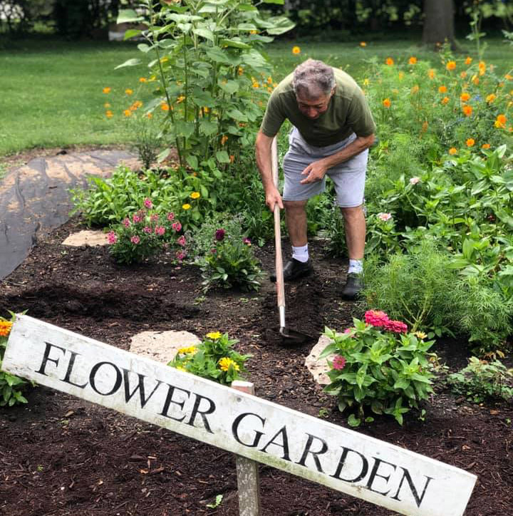 A man uses a garden hoe to cultivate the soil of a flower garden. In the foreground is a sign that reads "Flower Garden"