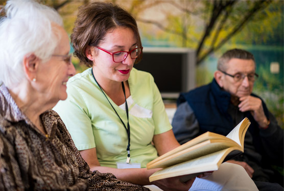 A woman in red glasses and a light green shirt reads from a book to an elderly woman sitting next to her. Some ways down, an elderly man looks off in a different direction.