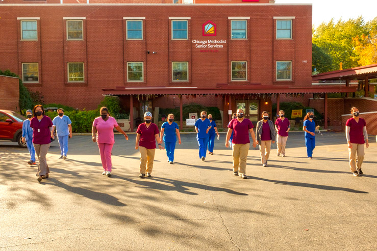 A large group of nurses, half in red and tan scrubs and half in all blue scrubs, engage in a walking exercise in the parking lot outside of the Chicago Methodist Senior Services building