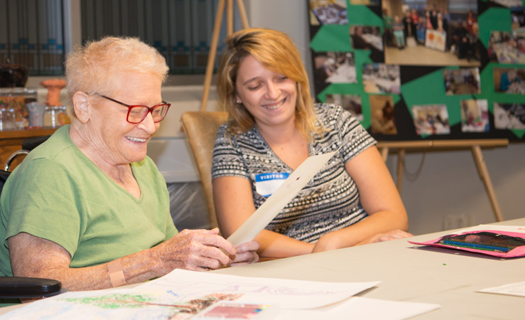An elderly woman and a young woman smile while looking over an art project