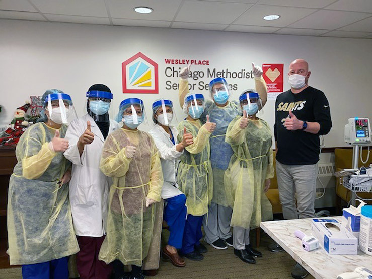 A group of medical professionals pose for a photo while adorned with face masks and face shields. Most, but not all, are also wearing yellow disposable medical gowns.