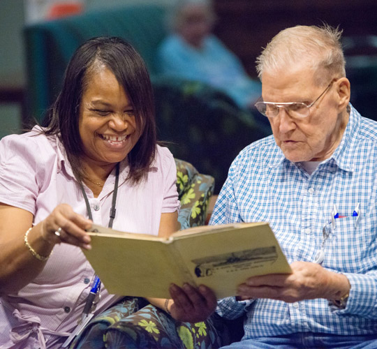 A Black woman in a pink shirt smiles while showing a book to a Caucasian elderly man in a blue plaid shirt.