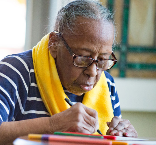 An elderly Black woman with a blue and white striped shirt and bright yellow scarf, writes in a notebook.