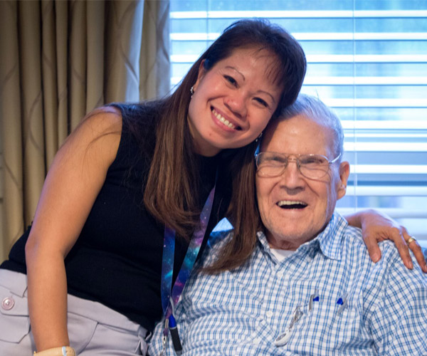 A young woman smiles as she hug an elderly man, who is also smiling.