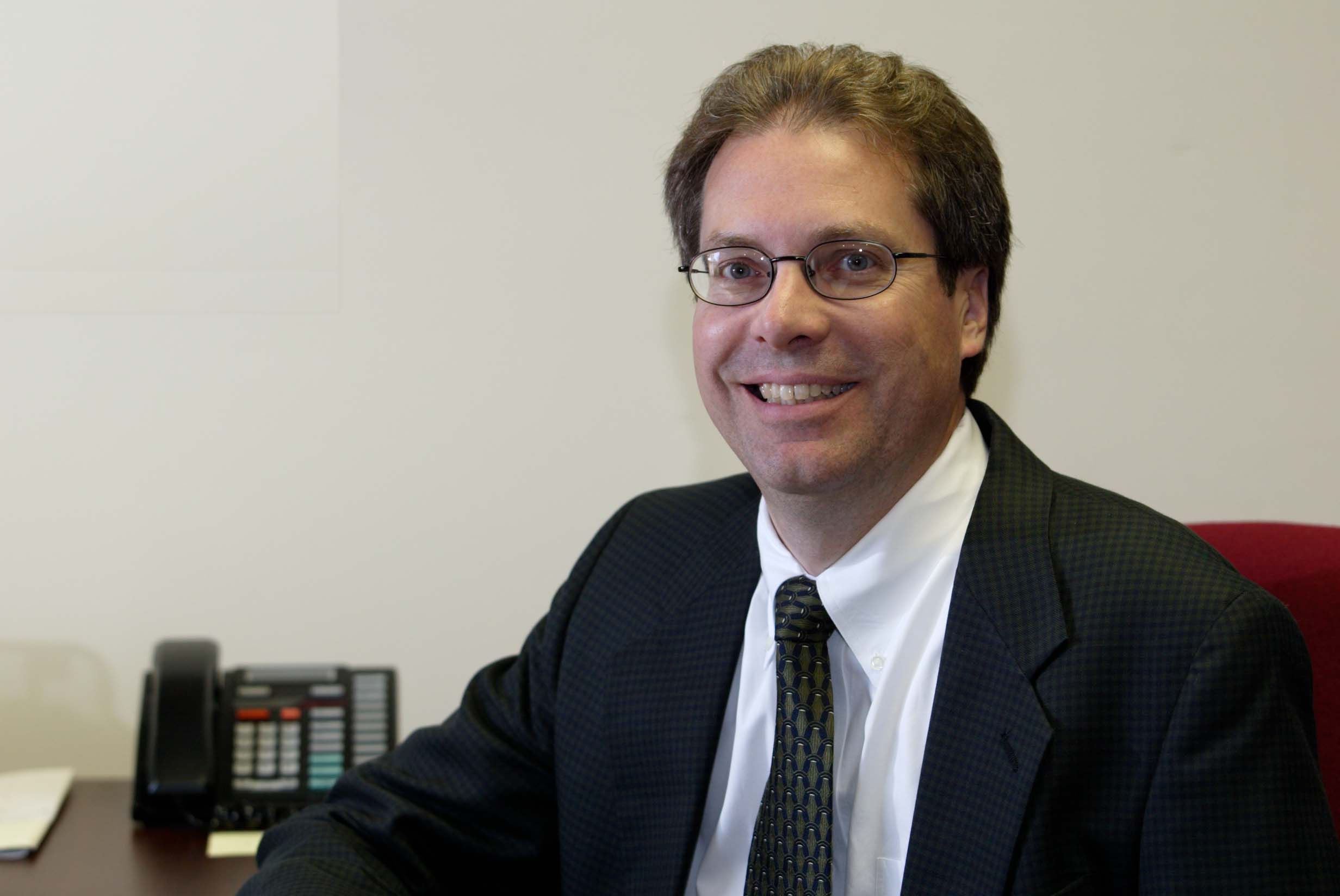 A man with short brown hair, glasses, and wearing a suit smiles and gazes just off from the camera.