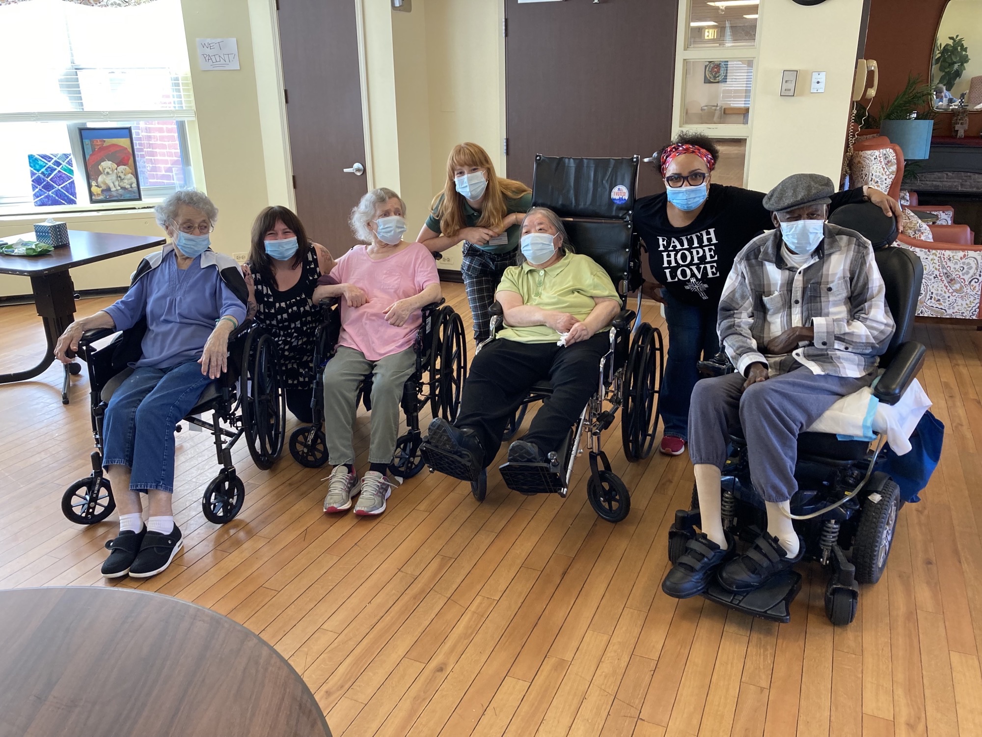 A group of seniors and their caregivers sit for a photo-op inside a care facility. All the seniors are seated in wheel chairs or motorized chairs. All people in the photo are wearing blue face masks.