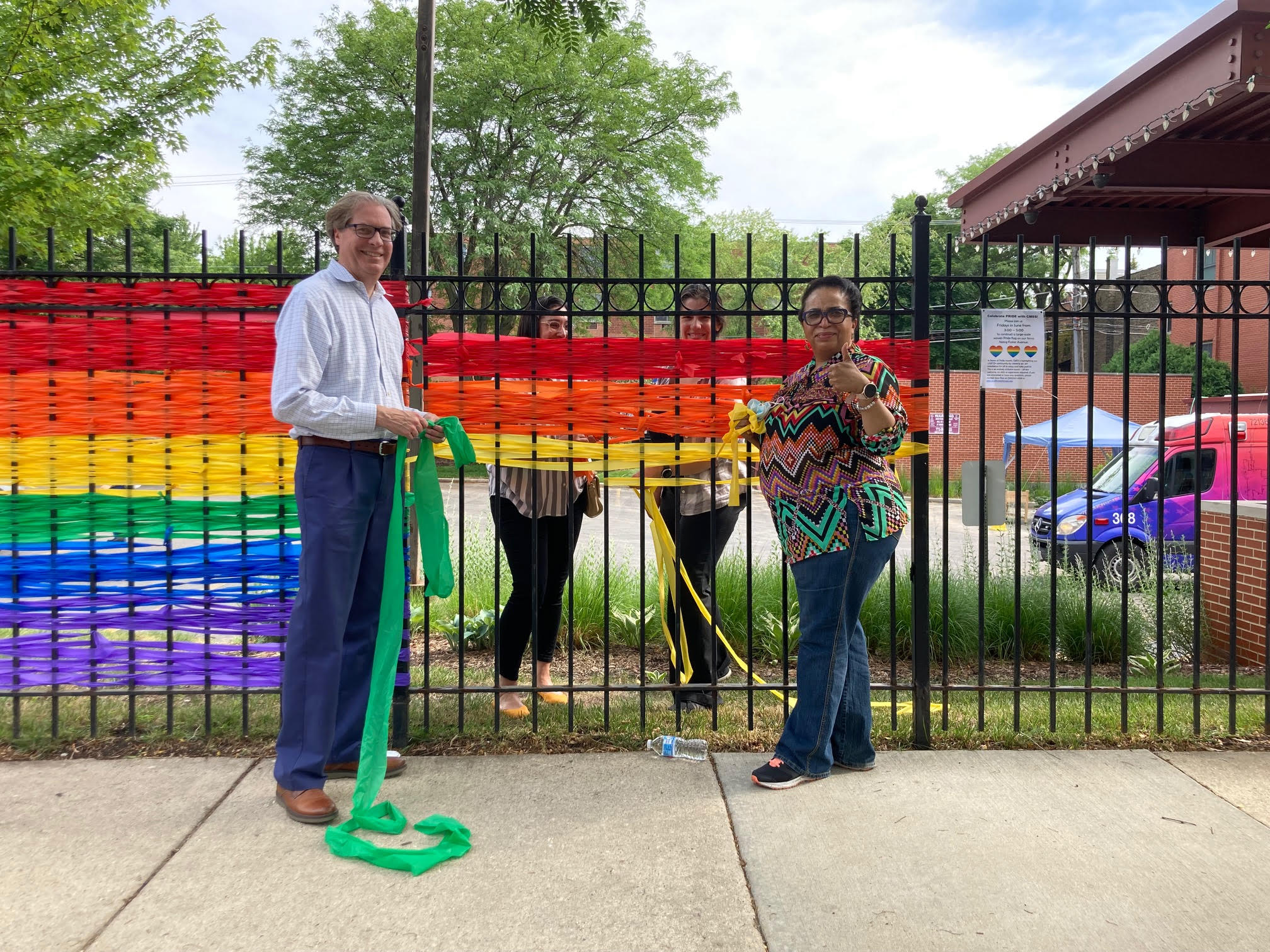 A man and woman put up ribbons in the color of the rainbow along an iron wrought fence in celebration of Pride.