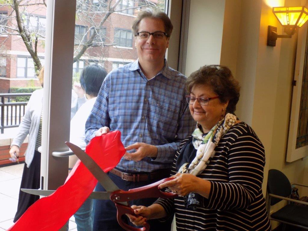 A man in a blue plaid shirt hold the end of a red ribbon. Beside him, a woman in a striped shirt holds an open pair of oversized scissors and is about to cut the ribbon.